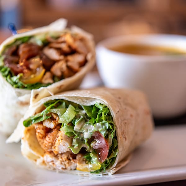 Two chicken wraps filled with lettuce, tomatoes, and sauce, served with a cup of soup.