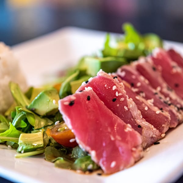 Seared tuna with sesame seeds, white rice, avocado, and greens on a white plate.