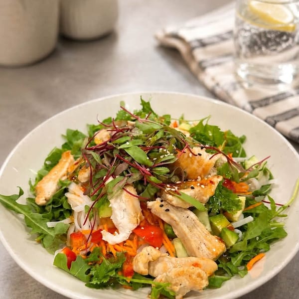A vibrant chicken salad with mixed greens, noodles, and vegetables in a white bowl.