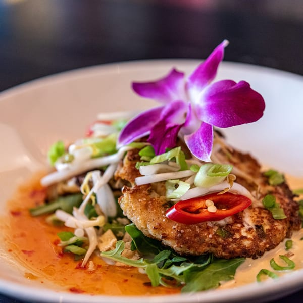 Two golden-brown crab cakes topped with bean sprouts, chili, and a purple orchid on a white plate.