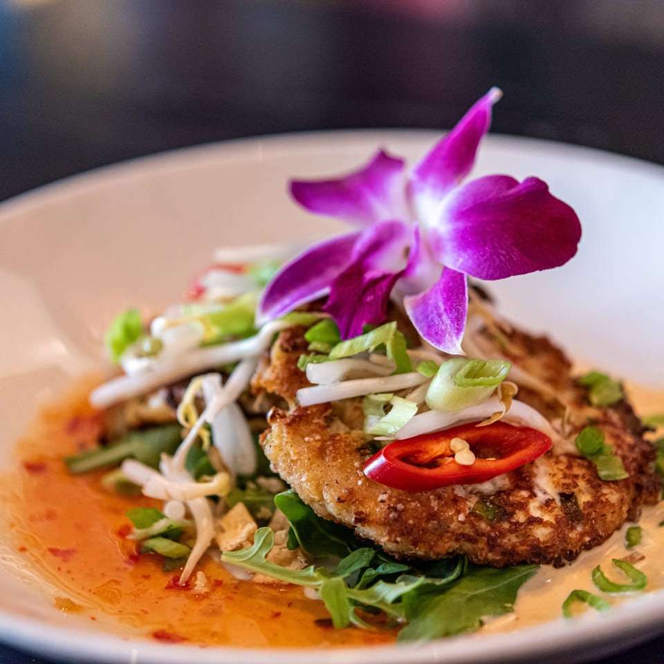 Two golden-brown crab cakes topped with bean sprouts, chili, and a purple orchid on a white plate.