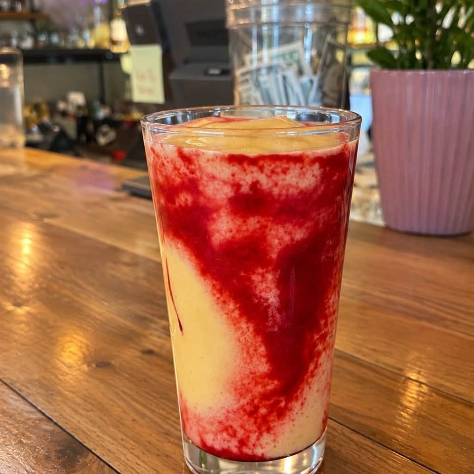 Red-and-cream layered smoothie topped with fruit sits on a wooden café counter beside a potted plant and liquor shelves.