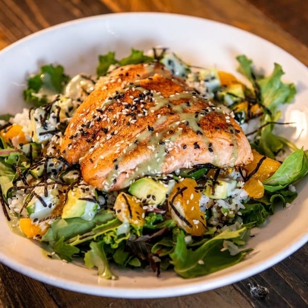 A salmon poke bowl with avocado, oranges, rice, and greens, drizzled with sauce.